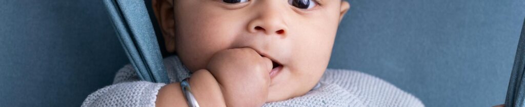 A cute baby dressed in blue, biting their thumb with a curious expression, against a matching blue background.